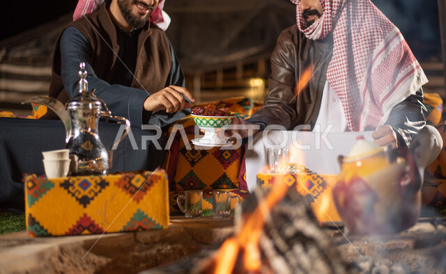 تقديم التمر للضيوف، مفهوم إكرام الضيوف، رجلان عربيان خليجيان سعوديان يرتديان الثوب التقليدي والشماغ مع ملابس الشتاء الدافئة يجلسان أمام شغلة النار، دلة قهوة عربية، وجبة ذات قيمة غذائية عالية، مخيمات سياحية في صحاري السعودية، الاستمتاع في قضاء الوقت مع الأصحاب