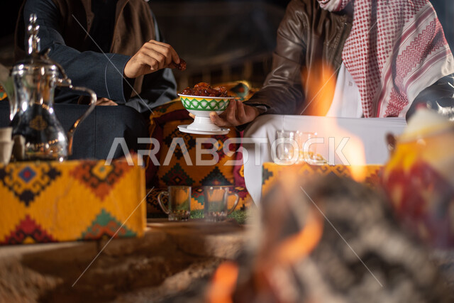 مفهوم إكرام الضيوف، رجلان عربيان خليجيان سعوديان يرتديان الثوب التقليدي والشماغ مع ملابس الشتاء الدافئة يجلسان أمام شغلة النار، تقديم التمر للضيوف، دلة قهوة عربية، وجبة ذات قيمة غذائية عالية، مخيمات سياحية في صحاري السعودية، الاستمتاع في قضاء الوقت مع الأصحاب