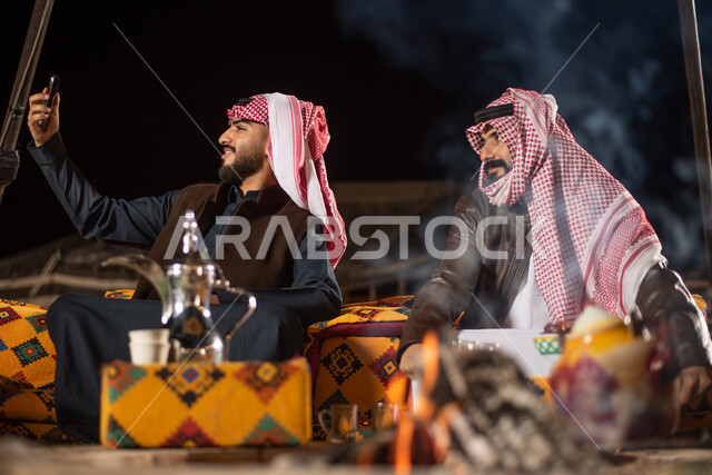 كشتة بر عربيه، إجراء مكالمات الفيديو والبثوث المباشرة عبر مواقع التواصل الاجتماعي، توثيق اللحظات السعيدة وقضاء أوقات ممتعة في الصحراء، رجلان عربيان خليجيان سعوديان يرتديان الثوب التقليدي مع ملابس الشتاء يقومان بالتقاط صورة سيلفي تذكرية عبر الهاتف المحمول