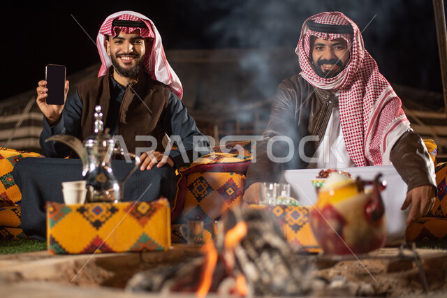 النظر إلى الكاميرا بإيماءات السرور، عرض شاشه سوداء فارغة عبر الجوال، شابان عربيان خليجيان سعوديان يرتديان الثوب التقليدي مع ملابس الشتاء يجلسون أمان النيران المشتعلة، استخدام التقنيات الحديثه المتطورة، مخيمات سياحية في الصحراء،  التدفئة من خلال شعلة النار
