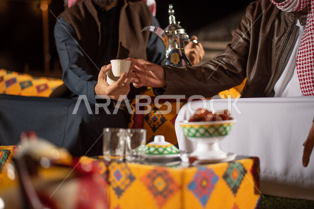 الكرم والضيافة عند العرب، تقديم المشروبات الساخنة، صب القهوة من الدلة النحاسية، صورة مقربة لرجلان عربيان خليجيان سعوديان يرتديان الثوب التقليدي مع ملابس الشتاء في رحلة تخييم برية، جلسة بر عربية شعبية، خيام شعبية في صحاري السعودية، قضاء أوقات ممتعة مع الأصدقاء