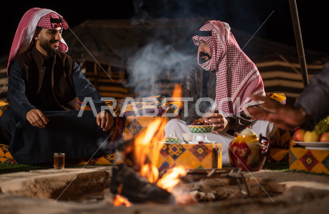 أجواء تخييم شبابية ليلية أمام شعلة الحطب، أصدقاء عربيون خليجيون سعوديون يجلسون أمام النيران المشتعلة في كشتة برية، جلسة بر عربية شعبية، مخيمات سياحية في صحاري السعودية، الاستمتاع في قضاء الوقت مع الأصحاب، ارتداء الملابس الشتوية الدافئة، حسن الضيافة والاستقبال