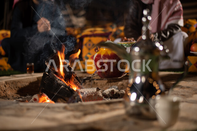 صديقان عربيان خليجيان سعوديان يرتديان الثوب التقليدي يجلسان امام شعلة النار، صورة مقربة لدلة القهوة العربية النحاسية مع فنجانين، مخيمات سياحية شعبية في صحاري السعودية، كشتة بر شبابية مسائية،  تقديم المشروبات الساخنة، مفهوم حسن الضيافة والاستقبال عند العرب