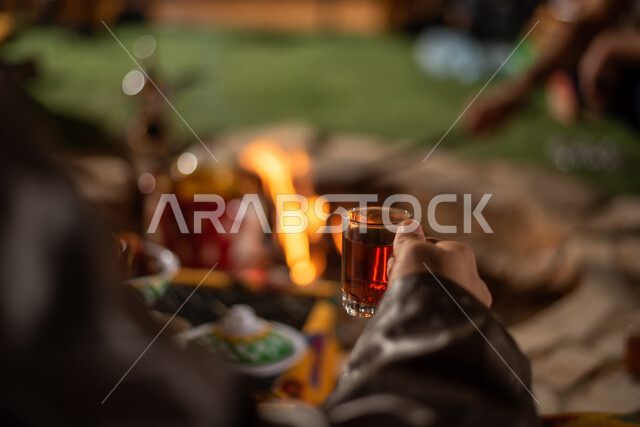 الجلسات الهادئة في البراري أمام موقد النار، الاستمتاع بالمشروبات الساخنة في الأجواء الشتوية، صورة مقربة لرجل سعودي عربي خليجي يستمتع بشرب الشاي، رحلة تخييم في صحاري السعودية، حسن الكرم والضيافه عند العرب، الكشتات البرية العربية الشعبية المسائية