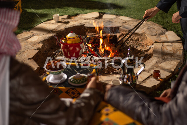 الاستمتاع بالمشروبات الساخنة، صورة مقربة لمجموعة من الاصدقاء العرب الخليجيين السعوديين يجلسون امام النبران المشتعلة، التدفئة من خلال شعلة النار، كشتة بر شبابية مسائية، الأجواء الهادئة في المخيمات الشعبيه في صحاري السعودية، التدفئة من خلال شعلة النار