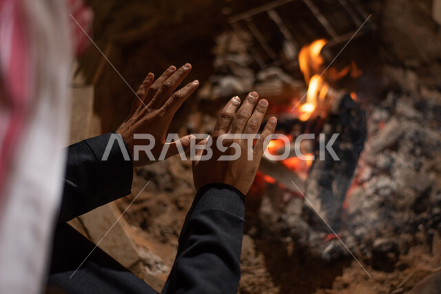 التدفئة من خلال شعلة النار، أجواء تخييم شبابية ليلية أمام شعلة الحطب، رجل عربي خليجي سعودي يرتدي الثوب التقليدي والشماغ يجلس أمام النيران المشتعلة في كشتة برية، جلسة بر عربية شعبية، مخيمات سياحية في صحاري السعودية