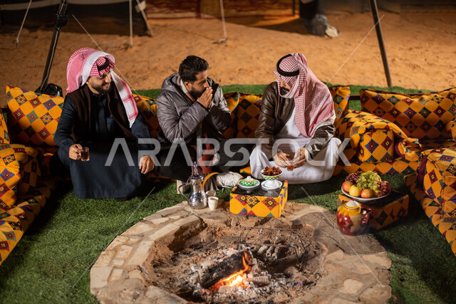 Good hospitality and reception, a youth camping atmosphere at night in front of a firewood flame, Saudi Gulf Arab friends sitting in front of the burning fires in a wild kashta, a popular Arab bar session, tourist camps in the deserts of Saudi Arabia, enjoying spending time with friends, wearing warm winter clothes.