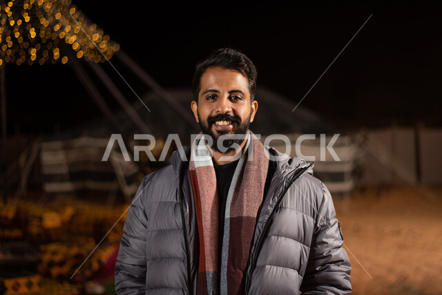 The concept of masculinity, elegance and elegance, attention to external appearance, a close-up image of a Saudi Gulf Arab man wearing a casual outfit with a thick gray winter jacket looking at the camera with gestures of happiness and pleasure, an evening youthful kashta, popular tourist camps in the deserts of Saudi Arabia, a night winter atmosphere