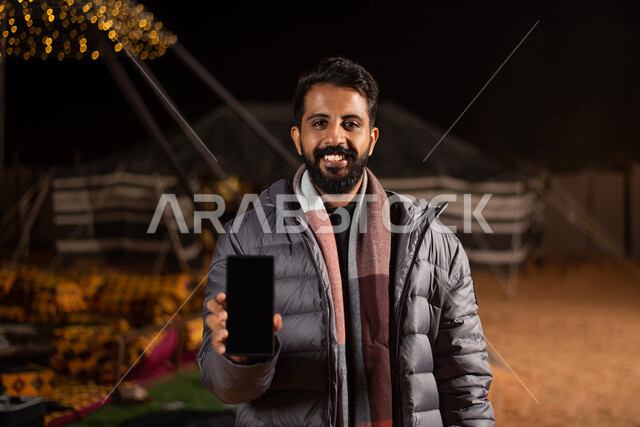 ايماءات السعادة والسرور، استخدام التقنيات الحديثة، عرض شاشة سوداء فارغة عبر الجوال، صورة مقربة لرجل عربي خليجي سعودي يرتدي الزي الكاجوال مع جاكيت رمادي شتوي سميك ينظر إلى الكاميرا، كشتة بر شبابية مسائية، مخيمات سياحية شعبية في صحاري السعودية، اجواء شتوية ليلية