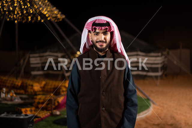 The concept of elegance and attention to external appearance, a close-up image of a Saudi Gulf Arab man wearing a traditional thobe with a brown leather jacket looking at the camera with gestures of happiness and pleasure, an evening youth camp, tourist camps in the desert, popular tents in the deserts of Saudi Arabia, a winter night atmosphere