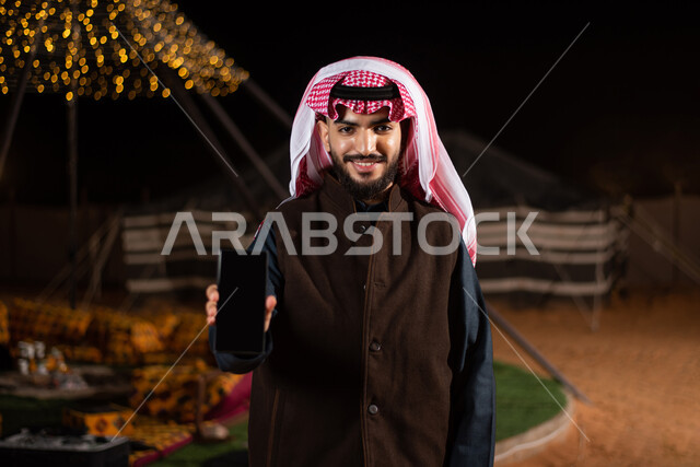 استخدام التقنيات الحديثة، عرض شاشة سوداء فارغة، صورة مقربة لرجل عربي خليجي سعودي يرتدي الثوب التقليدي والشماغ مع جاكيت بني جلد ينظر إلى الكاميرا يمسك بيده الهاتف المحمول، كشتة بر شبابية مسائية، مخيمات سياحية شعبية في صحاري السعودية، اجواء شتوية ليلية