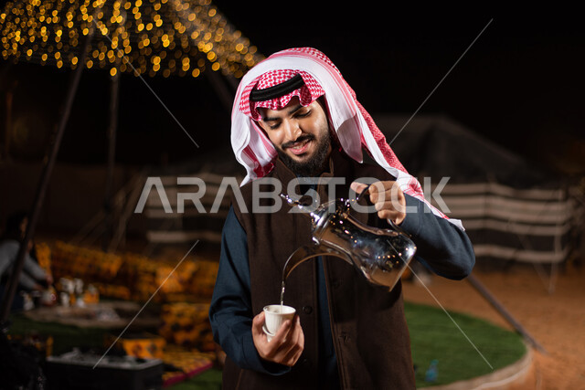 تقديم المشروبات الساخنة للضيوف، حسن الضيافة والاستقبال عند العرب، رحلة تخييم برية في الصحراء، صورة مقربة لرجل عربي خليجي سعودي يرتدي الثوب التقليدي مع ملابس الشتاء الدافئة يقوم بصب القهوة العربية، مخيمات سياحية في صحاري السعودية، كشتة بر عربية شبابية