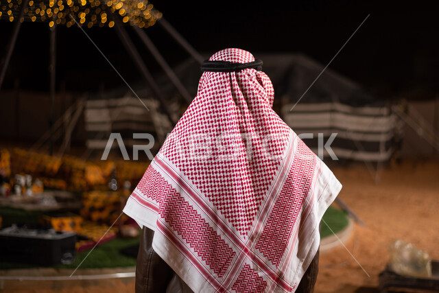 رحلة تخييم برية في المملكة، صورة من الخلف لرجل عربي خليجي سعودي يرتدي الثوب التقليدي والشماغ مع جاكيت جلد بني شتوي سميك، مخيمات سياحية شعبية في صحاري السعودية، كشتة بر شبابية مسائية، اجواء شتوية ليلية