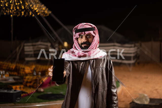 عرض شاشة سوداء فارغة، صورة مقربة لرجل عربي خليجي سعودي يرتدي الثوب التقليدي والشماغ مع جاكيت بني جلد يمسك بيده الهاتف المحمول، كشتة بر عربية شبابية مسائية، مخيمات سياحية شعبية في صحاري السعودية، اجواء شتوية ليلية، استخدام التقنيات الحديثة