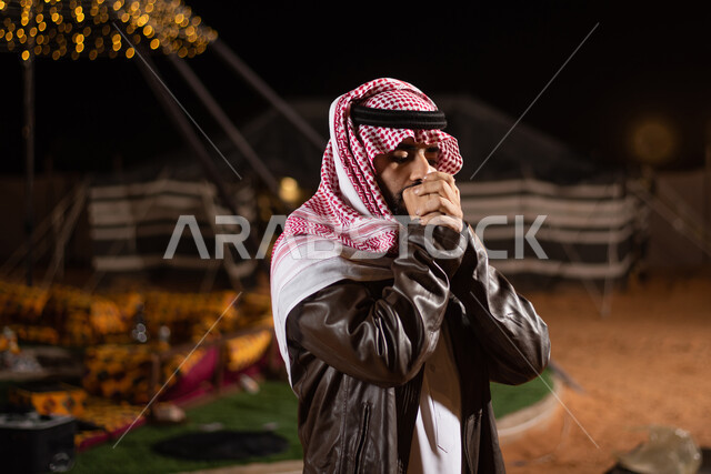 ايماءات التنفس على اليدين لابقائها دافئة، رحلة تخييم برية في المملكة، صورة مقربة لرجل عربي خليجي سعودي يرتدي الثوب التقليدي والشماغ مع ملابس الشتاء، جاكيت جلد بني شتوي سميك، مخيمات سياحية في الصحراء، خيام شعبية في صحاري السعودية، اجواء شتوية ليلية