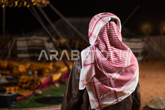 كشتة بر شبابية مسائية، رحلة تخييم برية في المملكة، صورة من الخلف لرجل عربي خليجي سعودي يرتدي الثوب التقليدي والشماغ مع جاكيت جلد بني شتوي سميك، مخيمات سياحية شعبية في صحاري السعودية، اجواء شتوية ليلية