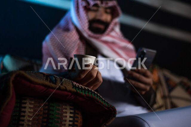 Drinking hot coffee, tourist camps in the desert, using modern and advanced technologies, browsing websites and social networking, chatting and talking with friends online, a Saudi Gulf Arab man wearing a traditional thobe and winter clothes, using a mobile phone.