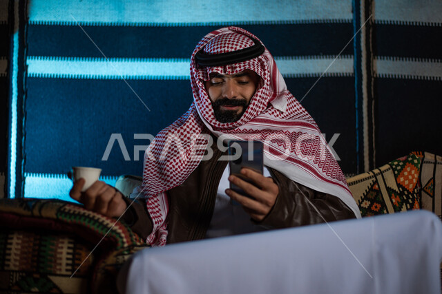 استخدام التقنيات الحديثة والمتطورة، تصفح المواقع الالكترونية والتواصل الاجتماعي، شرب القهوة الساخنة، مخيمات سياحية في الصحراء، الدردشة والتحدث مع الاصدقاء عبر الانترنت، رجل عربي خليجي سعودي يرتدي الثوب التقليدي مع ملابس الشتاء يستخدم الهاتف المحمول