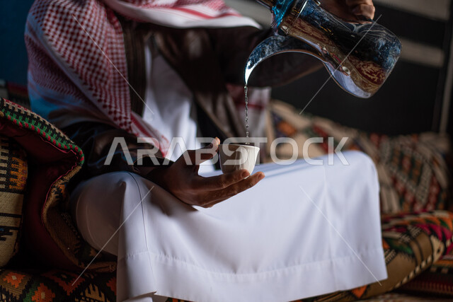 حسن الضيافة والاستقبال عند العرب، تقديم المشروبات الساخنة للضيوف، صورة مقربة ليد رجل عربي خليجي سعودي يرتدي ملابس الشتاء الدافئة والشماغ يقوم يصب القهوة، مخيمات سياحية في الصحراء، كشتة برعربية شبابية، اجواء شتوية ليلية،  خيام شعبية في صحاري السعودية