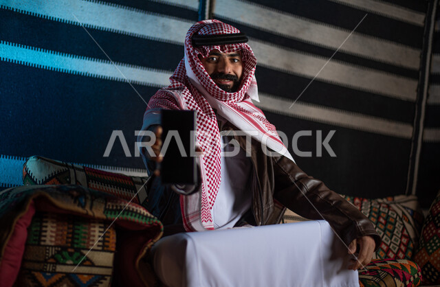 عرض خدمات وتطبيقات الجوال، استخدام التقنيات الحديثه المتطورة، صورة مقربة لرجل عربي خليجي سعودي يرتدي ملابس الشتاء الدافئة والشماغ يمسك بيده هاتف محمول بشاشة سوداء فارغة، مخيمات سياحية في الصحراء، كشتة برعربية شبابية، اجواء شتوية ليلية