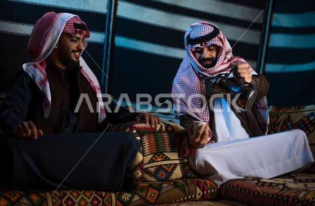 Pouring coffee from a copper pot, spending enjoyable times with friends, good hospitality and reception, two Saudi Gulf Arab men wearing traditional thobes and shemaghs with winter clothes on a wild camping trip in the desert, a popular Arab camping session, popular tents in the deserts of Saudi Arabia, a winter night atmosphere