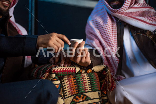 إكرام الضيف و حسن الضيافه والاستقبال، حوارات ونقاشات بين الاصدقاء، تقديم القهوة العربية للضيوف، الاستمتاع بشرب المشروبات الساخنة، شابان عربيان خليجيان سعوديان يرتديان الثوب التقليدي مع ملابس الشتاء في رحلة تخييم برية في صحراء السعودية، كشتة بر شبابية مسائية