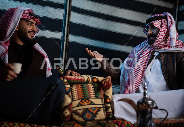 الاستمتاع بشرب المشروبات الساخنة، تقديم القهوة العربية للضيوف، حسن الضيافه والكرم، شابان عربيان خليجيان سعوديان يرتديان الثوب التقليدي مع ملابس الشتاء في رحلة تخييم برية في صحراء السعودية، حوارات ونقاشات بين الاصدقاء، كشتة بر شبابية مسائية، اجواء شتوية ليلية