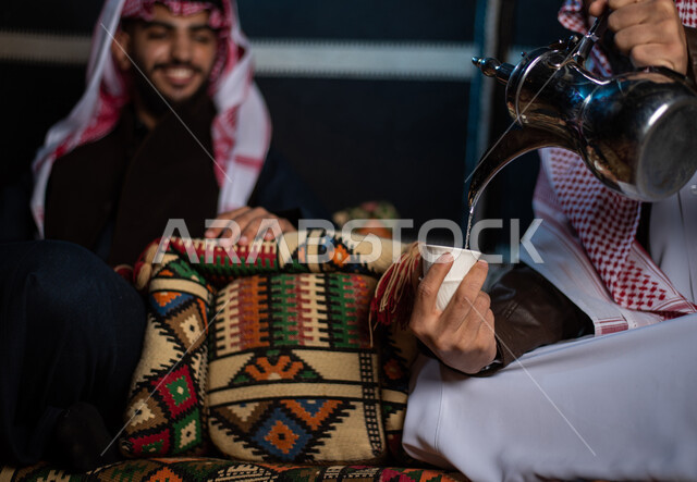 تقديم المشروبات الساخنة للأصدقاء، ايماءات البشاشة والترحيب، صب القهوة من الدلة النحاسية، رجلان عربيان خليجيان سعوديان يرتديان الثوب التقليدي مع ملابس الشتاء في رحلة تخييم برية في الصحراء، جلسة برعربية شعبية، خيام شعبية في صحاري السعودية، أجواء شتوية ليلية