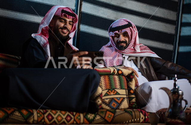 الجلسات السعودية التقليدية الشعبية، قضاء أوقات ممتعة في الأجواء الليلية الشتوية في الصحراء، إجراء مكالمات الفيديو والبثوث المباشرة عبر مواقع التواصل الاجتماعي، رجلان عربيان خليجيان سعوديان يرتديان الثوب التقليدي مع ملابس الشتاء الدافئة يستخدمان الهاتف المحمول
