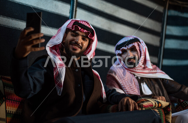 كشتة بر عربيه، توثيق اللحظات السعيدة وقضاء أوقات ممتعة في الصحراء، إجراء مكالمات الفيديو والبثوث المباشرة عبر مواقع التواصل الاجتماعي، رجلان عربيان خليجيان سعوديان يرتديان الثوب التقليدي مع ملابس الشتاء يقومان بالتقاط صورة سيلفي تذكرية عبر الهاتف المحمول