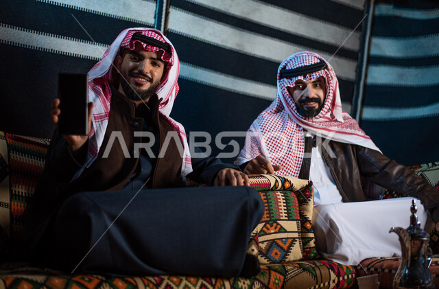 The use of advanced modern technologies, tourist camps in the desert, a youthful Berber Kashta, a winter night atmosphere, a blank black screen shown, two young Saudi Gulf Arab men wearing the traditional thobe and shemagh with warm winter clothes looking at the camera with gestures of pleasure and happiness.