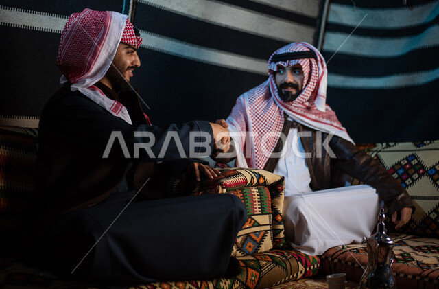 الترحيب والسلام وإلقاء التحية، رجلان عربيان خليجيان سعوديان يرتديان الثوب التقليدي والشماغ مع ملابس الشتاء الدافئة يتصافحان بإيماءات السرور، مخيمات سياحية في الصحراء، كشتة بر عربية شبابية، قضاء أوقات ممتعة مع الأصدقاء، الجلسات السعودية التقليدية الشعبية