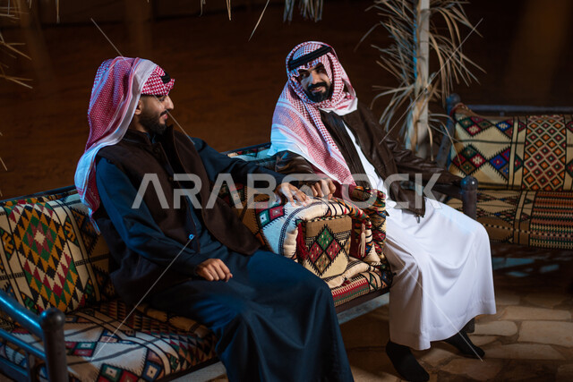 مخيمات سياحية في المناطق البرية، حوارات ونقاشات بين الاصدقاء، رجلان عربيان خليجيان سعوديان يرتديان الثوب التقليدي والشماغ مع ملابس الشتاء الدافئة يقضون أوقات ممتعة في رحلة تخييم في صحاري السعودية، خيام شعبية في الصحراء، كشتة بر شبابية، اجواء شتوية ليلية