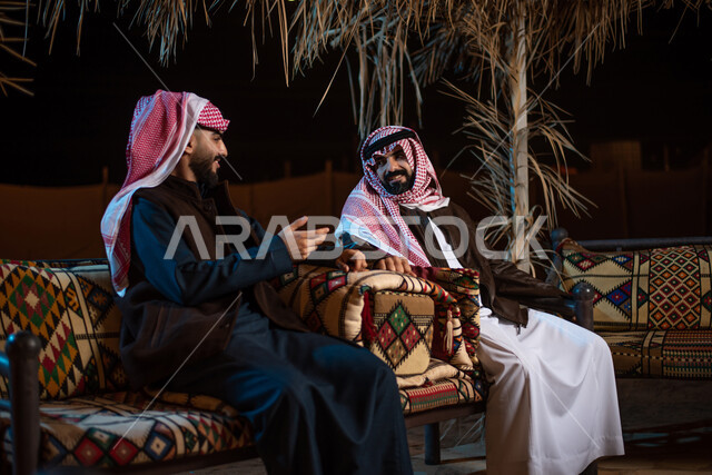 حوارات ونقاشات بين الاصدقاء، رجلان عربيان خليجيان سعوديان يرتديان الثوب التقليدي والشماغ مع ملابس الشتاء الدافئة يقضون أوقات ممتعة في رحلة تخييم في صحاري السعودية، خيام شعبية في الصحراء، كشتة بر شبابية، اجواء شتوية ليلية، مخيمات سياحية في المناطق البرية