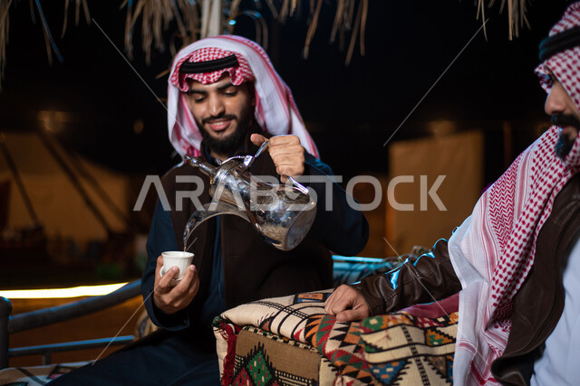Pouring coffee from a copper pot, good hospitality and reception, two Saudi Gulf Arab men wearing the traditional thobe and shemagh with winter clothes on a wild camping trip in the desert, a popular Arab camping session, popular tents in the deserts of Saudi Arabia, a winter night atmosphere, spending fun times with friends