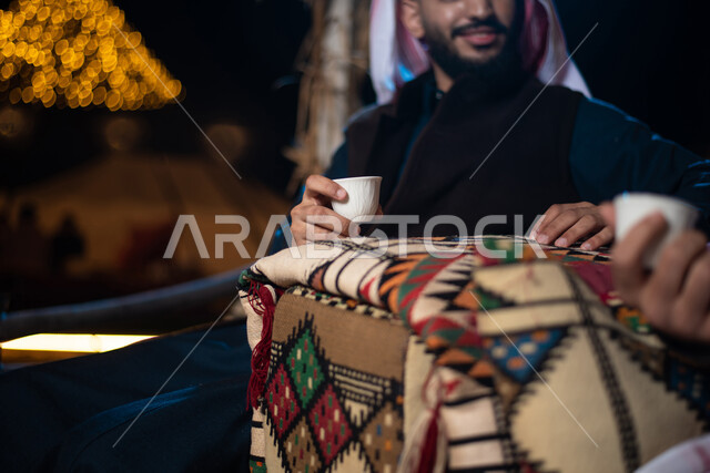تقديم القهوة العربية الساخنة للضيوف، مفهوم حسن الضيافه والكرم، شابان عربيان خليجيان سعوديان يرتديان الثوب التقليدي والشماغ مع ملابس الشتاء في رحلة تخييم برية في صحراء السعودية، حوارات ونقاشات بين الاصدقاء، كشتة بر شبابية مسائية، اجواء شتوية ليلية