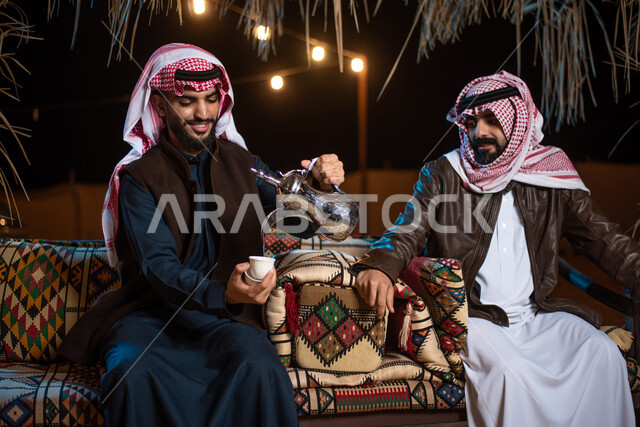 إيماءات البشاشة والترحيب، حسن الضيافة والإستقبال، صب القهوة من الدلة النحاسية، رجلان عربيان خليجيان سعوديان يرتديان الثوب التقليدي مع ملابس الشتاء في رحلة تخييم برية في الصحراء، جلسة بر عربية شعبية، خيام شعبية في صحاري السعودية،  قضاء أوقات ممتعة  مع الأصدقاء