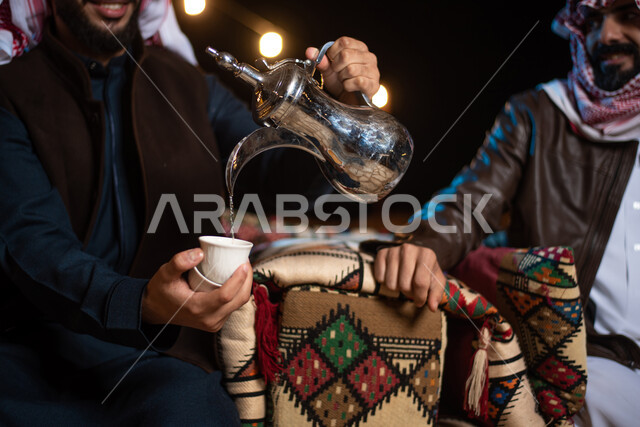 حسن الضيافة والإستقبال، صب القهوة من الدلة النحاسية، رجلان عربيان خليجيان سعوديان يرتديان الثوب التقليدي والشماغ مع ملابس الشتاء في رحلة تخييم برية في الصحراء، جلسة بر عربية شعبية، خيام شعبية في صحاري السعودية، أجواء ليلية شتوية،  قضاء أوقات ممتعة  مع الأصدقاء