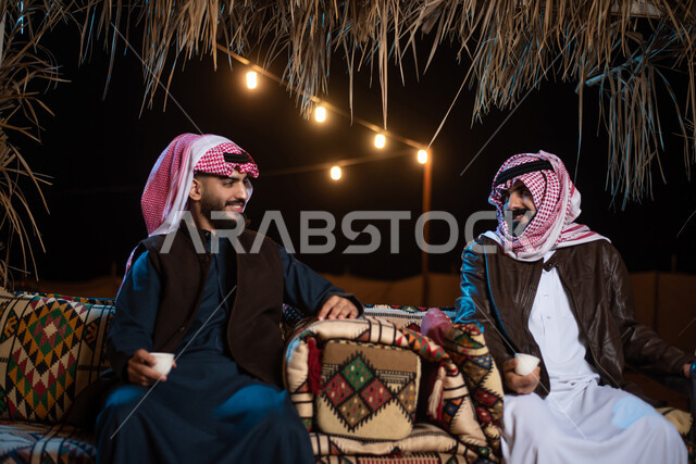 مفهوم حسن الضيافه والكرم، شابان عربيان خليجيان سعوديان يرتديان الثوب التقليدي والشماغ مع ملابس الشتاء في رحلة تخييم برية في صحراء السعودية، تقديم القهوة العربية الساخنة للضيوف، حوارات ونقاشات بين الاصدقاء، كشتة بر شبابية مسائية، اجواء شتوية ليلية