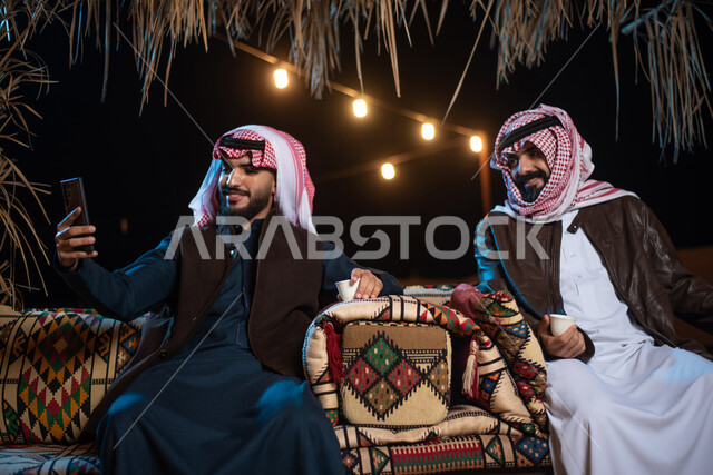 Making video calls and live broadcasts via social networking sites, documenting happy moments and spending enjoyable times in the desert, two Saudi Gulf Arab men wearing traditional thobes and winter clothes taking a memorable selfie via mobile phone, Kashta Bar Arabia