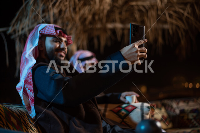 توثيق اللحظات السعيدة وقضاء أوقات ممتعة في الصحراء، إجراء مكالمات الفيديو والبثوث المباشرة عبر مواقع التواصل الاجتماعي، رجلان عربيان خليجيان سعوديان يرتديان الثوب التقليدي مع ملابس الشتاء يقومان بالتقاط صورة سيلفي تذكرية عبر الهاتف المحمول، كشتة بر عربيه