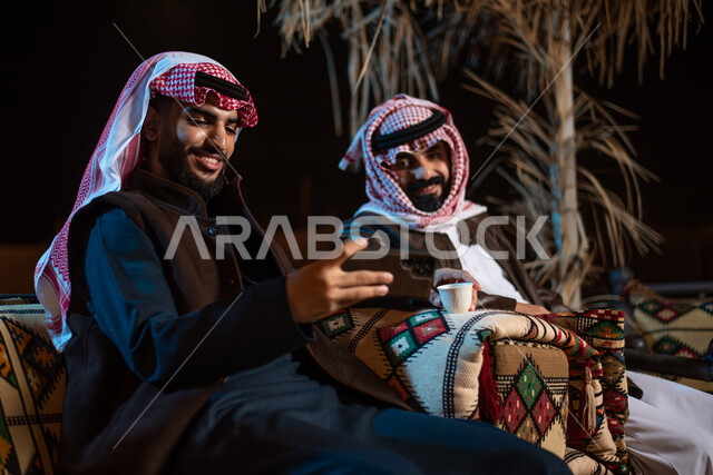 قضاء أوقات ممتعة في الأجواء الليلية الشتوية في الصحراء، إجراء مكالمات الفيديو والبثوث المباشرة عبر مواقع التواصل الاجتماعي، رجلان عربيان خليجيان سعوديان يرتديان الثوب التقليدي مع ملابس الشتاء الدافئة يستخدمان الهاتف المحمول، الجلسات السعودية التقليدية الشعبية