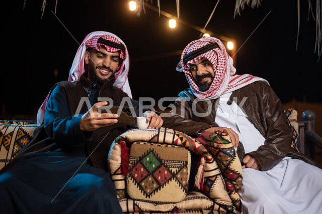 إجراء مكالمات الفيديو والبثوث المباشرة عبر مواقع التواصل الاجتماعي، رجلان عربيان خليجيان سعوديان يرتديان الثوب التقليدي مع ملابس الشتاء الدافئة يستخدمان الهاتف المحمول، الجلسات السعودية التقليدية الشعبية، قضاء أوقات ممتعة في الأجواء الليلية الشتوية في الصحراء