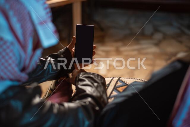 استخدام التقنيات الحديثه المتطورة، صورة مقربة ليد رجل عربي خليجي سعودي يرتدي ملابس الشتاء الدافئة والشماغ يمسك بيده هاتف محمول بشاشة سوداء فارغة، عرض خدمات وتطبيقات الجوال، مخيمات سياحية في الصحراء، كشتة برعربية شبابية، اجواء شتوية ليلية