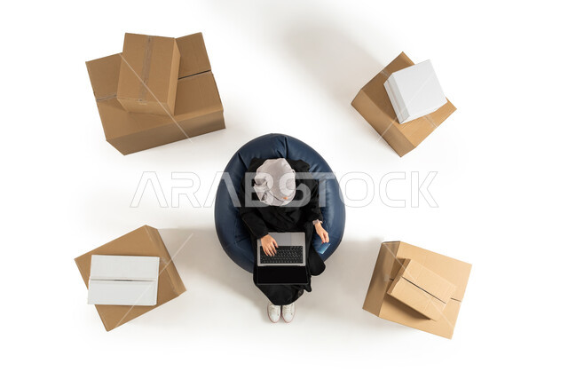 Distance shopping, sales and purchases, using a laptop, portrait of an overhead vertical image of a Saudi Arabian Gulf woman sitting on a chair, receiving the order box, online orders, white background