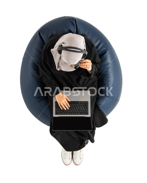 متابعة وتطوير الأعمال، العمل عن بعد، بورتريه لصورة علوية عامودية لشابة عربية خليجية سعودية تجلس على كرسي مريح تستخدم الحاسوب المحمول، شابة تضع سماعة الرأس ومستمتعة بسماع الاغاني والموسيقى، تسجيل الاغاني والصوتيات، انجاز مهام العمل،  دردشة وتواصل، خلفية بي