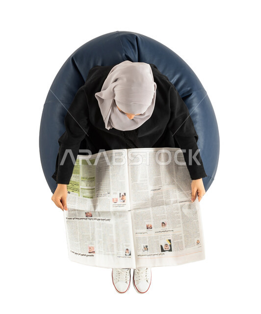 قضاء الوقت بالقراءه،  بورتريه لصورة علوية عامودية لسيدة عربية خليجية سعودية تجلس على كرسي مريح وتحمل بيدها جريدة ورقية ، التعلم والثقافة ، ثقافة القراءة، المعرفة، خلفية بيضاء