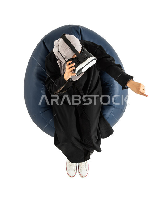 Leisure and entertainment, 3D glasses, enjoying watching movies, portrait of a vertical overhead image of a Saudi Arabian Gulf woman sitting on a comfortable chair and wearing virtual reality glasses holding a joystick in her hand and playing video games,