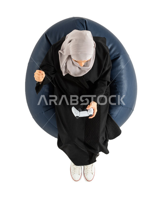 المتعة و التسلية بألعاب الفيديو، بورتريه لصورة علوية عامودية لسيدة عربية خليجية سعودية تجلس على كرسي مريح، تمسك بيدها عصا التحكم و تلعب بألعاب الفيديو،  ألعاب البلايستيشن الترفيهية، إيماءات تدل على الفوز والسعادة ، تقنيات و ألعاب, خلفية بيضاء