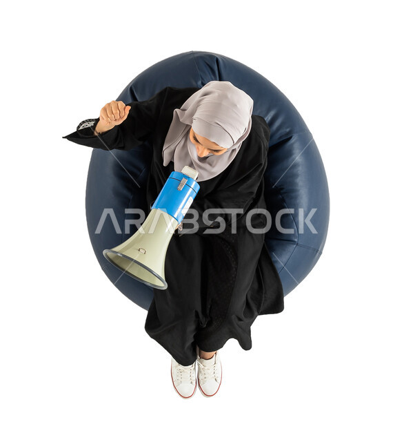 Using the sound horn, an overhead vertical image of a Saudi Arab Gulf woman sitting on a comfortable chair, holding a loudspeaker in her hand, white background